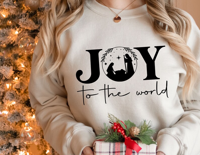 Pu&ograve; includere: Una felpa bianca con le parole "JOY to the world" stampate in nero. La parola "JOY" &egrave; stilizzata con un cerchio che contiene una silhouette nera della scena della Nativit&agrave;.
