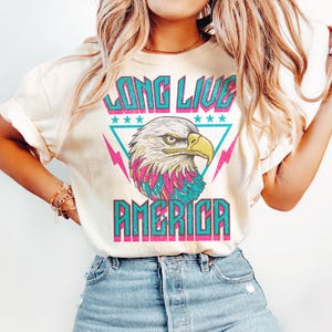 May include: A white t-shirt with a vintage-style graphic of an eagle with the text "Long Live America" in turquoise, pink, and white.