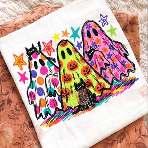 K&ouml;nnte beinhalten: Ein wei&szlig;es T-Shirt mit einer bunten Grafik von drei Geistern und drei schwarzen Katzen. Die Geister sind mit Punkten, K&uuml;rbissen und Blumen verziert. Das Design enth&auml;lt leuchtende Sterne in Gelb, Orange, Blau und Lila sowie eine regenbogenfarbene Basis.