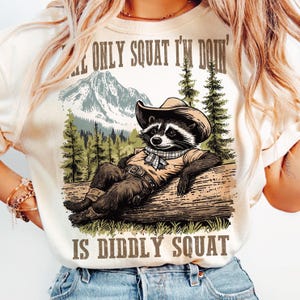 May include: Cream-colored t-shirt with a graphic of a raccoon wearing a cowboy hat, resting on a log. The raccoon is in front of a mountain and forest scene. Text on the shirt reads, "THE ONLY SQUAT I'M DOIN' IS DIDDLY SQUAT."