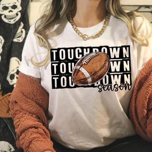 Op de afbeelding: Wit T-shirt met de tekst "Touchdown Season" en een afbeelding van een bruine en witte voetbal.