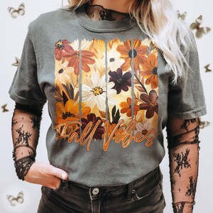 May include: Gray t-shirt with a floral design in shades of orange, brown, and white. The words "Fall Vibes" are printed in orange script across the bottom of the floral arrangement. The shirt has short sleeves.