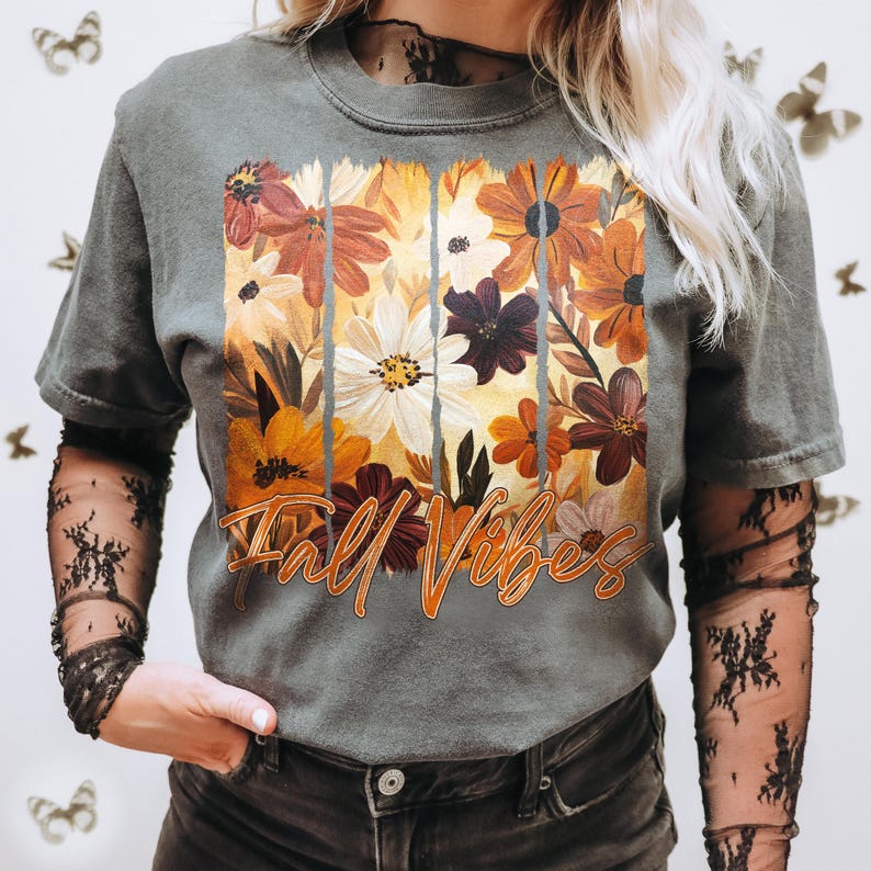May include: Gray t-shirt with a floral design in shades of orange, brown, and white. The words "Fall Vibes" are printed in orange script across the bottom of the floral arrangement. The shirt has short sleeves.