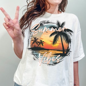 May include: White t-shirt with a graphic print of a sunset over the ocean with palm trees and the text "Summer Vibes".