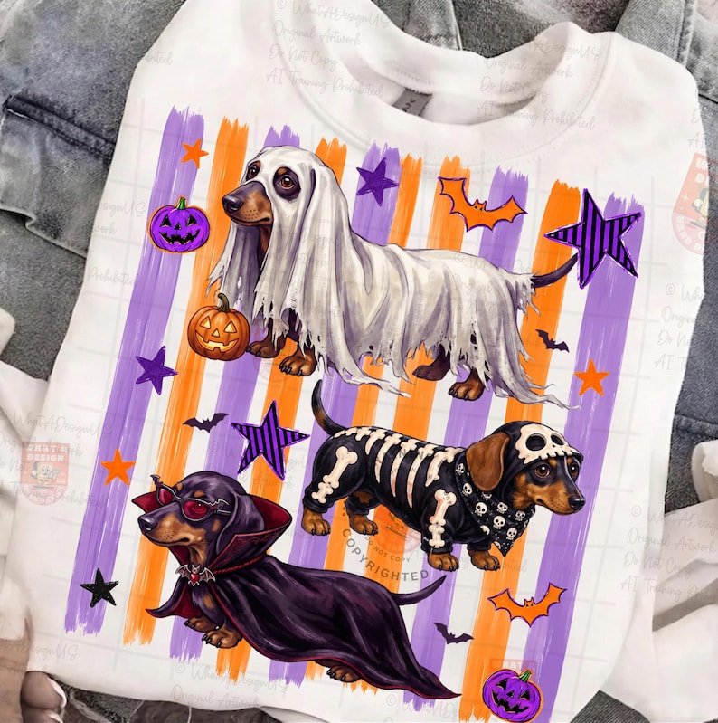 May include: A white sweatshirt featuring a graphic print of three dachshunds in Halloween costumes: one as a ghost, one as a skeleton, and one as a vampire. The background includes vertical orange and purple stripes, orange and purple stars, bats, and purple jack-o'-lanterns.