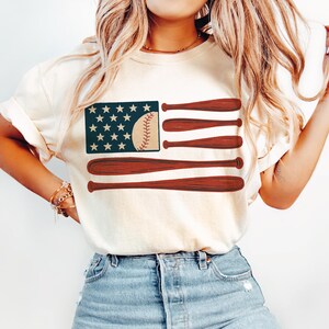 May include: Cream-colored t-shirt featuring a baseball-themed American flag design. The flag's stars are represented by white stars on a navy blue background. The stripes are replaced with brown baseball bats and a baseball.