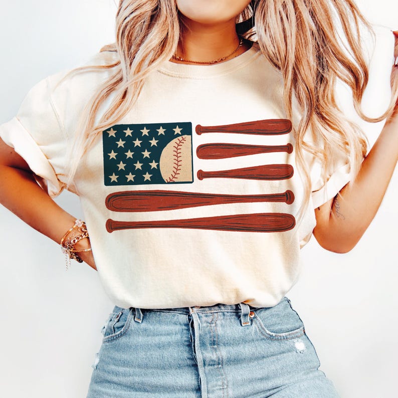 May include: Cream-colored t-shirt featuring a baseball-themed American flag design. The flag's stars are represented by white stars on a navy blue background. The stripes are replaced with brown baseball bats and a baseball.