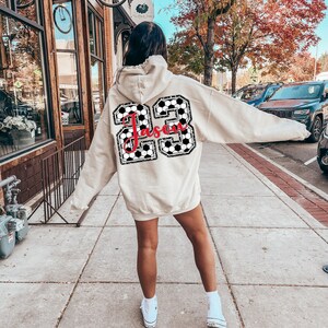 May include: A beige hoodie with a black and white checkered soccer ball design and the name "Jason" in red lettering. The hoodie is worn by a person walking down a sidewalk.