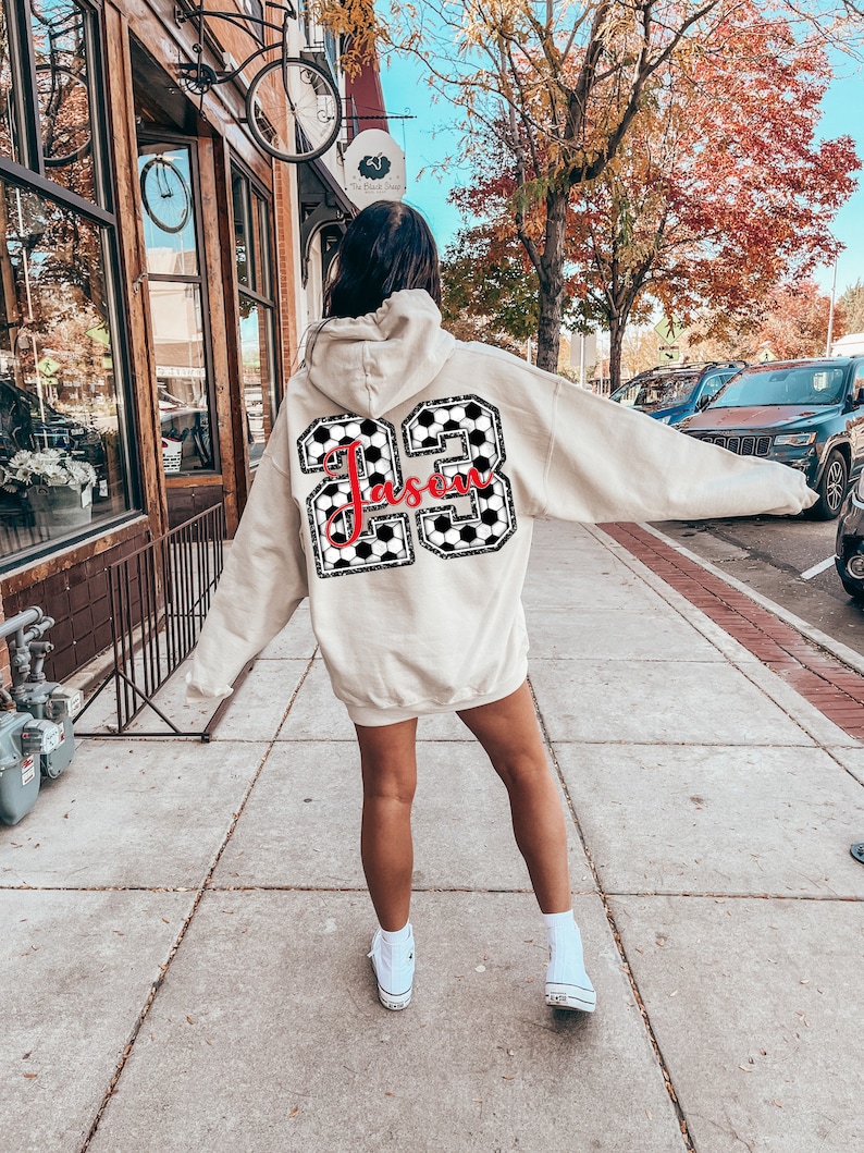 May include: A beige hoodie with a black and white checkered soccer ball design and the name "Jason" in red lettering. The hoodie is worn by a person walking down a sidewalk.