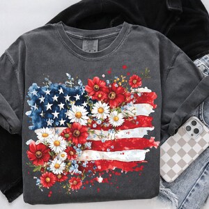 May include: A dark gray t-shirt with a graphic of an American flag surrounded by red, white, and blue flowers. The flag is painted in a watercolor style with white splatters.