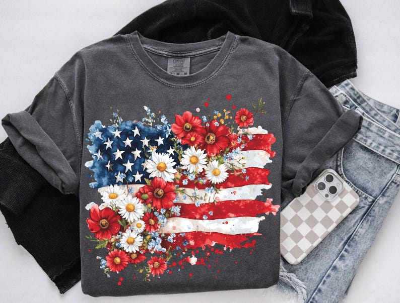 May include: A dark gray t-shirt with a graphic of an American flag surrounded by red, white, and blue flowers. The flag is painted in a watercolor style with white splatters.