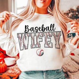May include: White sweatshirt with the text "Baseball Wife" in black letters with a baseball stitch design. A baseball is below the text.