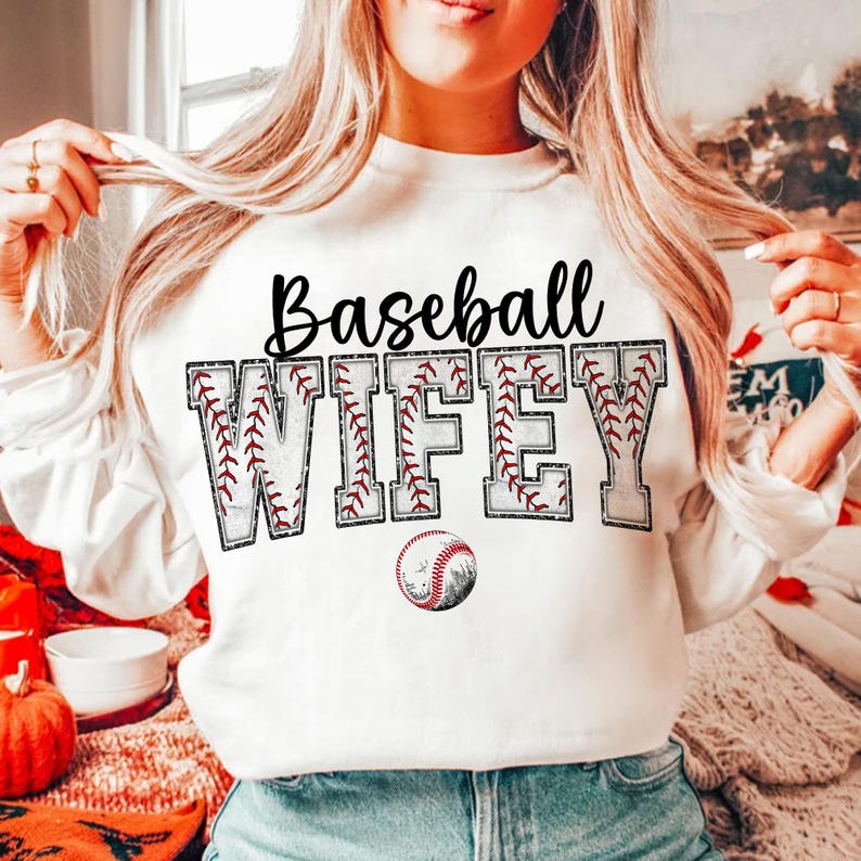 May include: White sweatshirt with the text "Baseball Wife" in black letters with a baseball stitch design. A baseball is below the text.