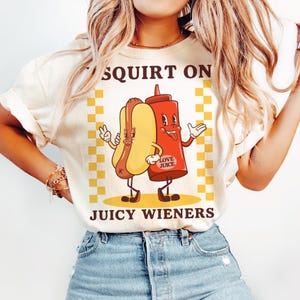 May include: A white t-shirt with a graphic of a hot dog and ketchup bottle with the text "SQUIRT ON JUICY WIENERS" and the words "LOVE JUICE" on the ketchup bottle.