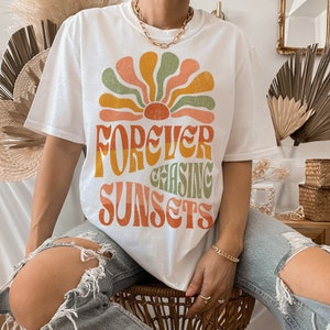 May include: White t-shirt with a retro-style graphic of a sunburst design in orange, yellow, green, and peach. The text "FOREVER CHASING SUNSETS" is printed below the sunburst. The shirt is a casual style.