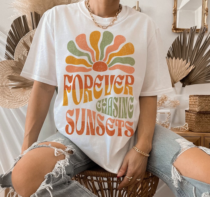 May include: White t-shirt with a retro-style graphic of a sunburst design in orange, yellow, green, and peach. The text "FOREVER CHASING SUNSETS" is printed below the sunburst. The shirt is a casual style.