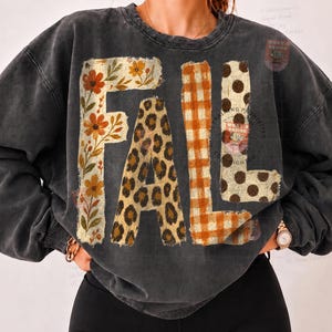 May include: A charcoal grey crewneck sweatshirt featuring the word FALL in large block letters. Each letter has a distinct pattern: F has orange and yellow flowers, A has a leopard print, L has an orange gingham check, and L has brown polka dots.
