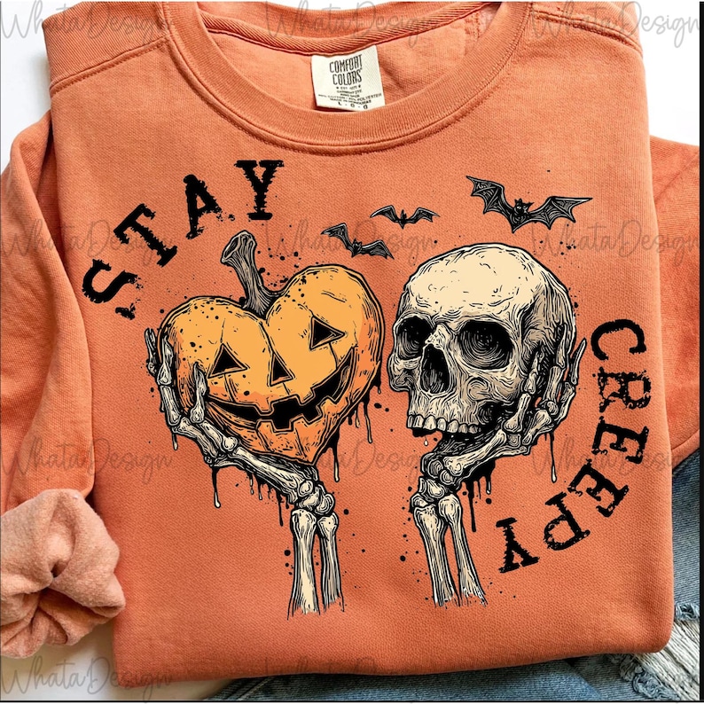 Digital Stay Creepy Halloween PNG design featuring a skeleton hand dripping a heart, with bats and a moon in a retro grunge style. Shown on an orange t-shirt mockup.