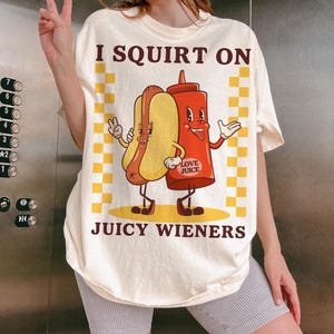 May include: A white t-shirt with a graphic of a hot dog and a ketchup bottle with the text "I SQUIRT ON JUICY WIENERS" and "LOVE JUICE" on the bottle.