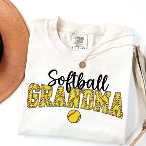 May include: A white t-shirt with the text "Softball Grandma" in black lettering. The letters are outlined in yellow and have a softball stitch pattern. A yellow softball is below the text.