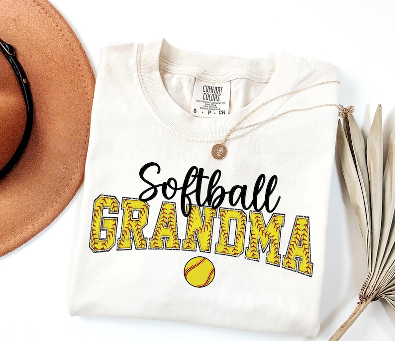 May include: A white t-shirt with the text "Softball Grandma" in black lettering. The letters are outlined in yellow and have a softball stitch pattern. A yellow softball is below the text.