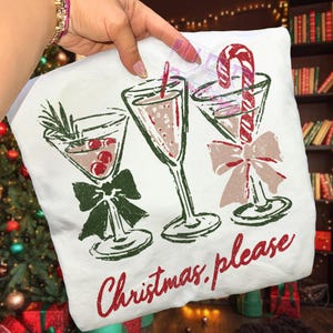 May include: White t-shirt with a festive design featuring three martini glasses with holiday garnishes and bows. The text "Christmas please" is printed in red script below the glasses.