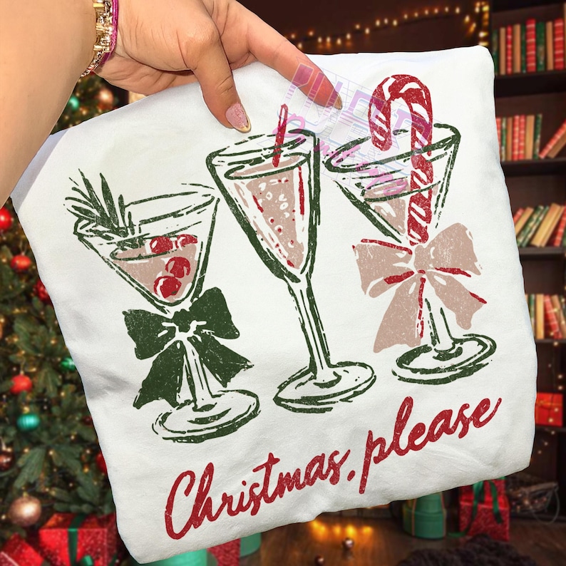 May include: White t-shirt with a festive design featuring three martini glasses with holiday garnishes and bows. The text "Christmas please" is printed in red script below the glasses.