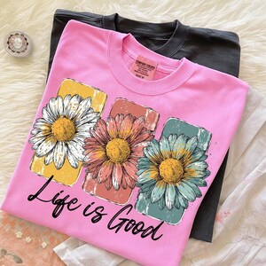 May include: A pink t-shirt with a daisy graphic and the text "Life is Good." The graphic features three daisies in yellow, peach, and blue rectangles. A dark gray t-shirt is visible underneath.