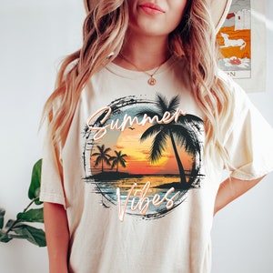 May include: A cream colored t-shirt with a graphic of a sunset over a beach with palm trees and the text "Summer Vibes" in a distressed font.