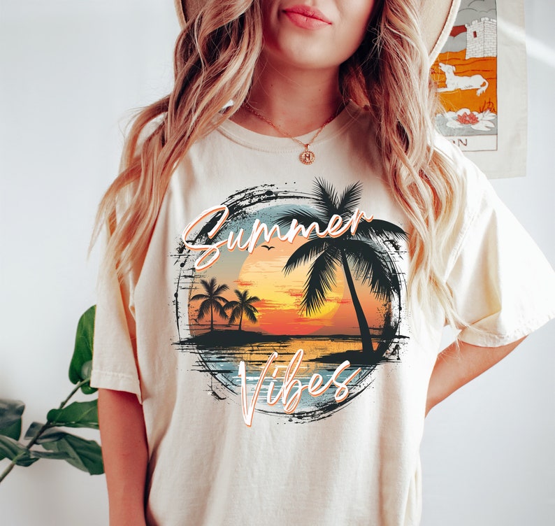 May include: A cream colored t-shirt with a graphic of a sunset over a beach with palm trees and the text "Summer Vibes" in a distressed font.
