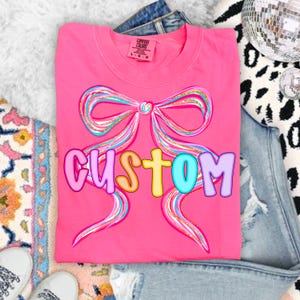 K&ouml;nnte beinhalten: Ein leuchtend pinkfarbenes T-Shirt mit einem gro&szlig;en Bogen-Design in Blau-, Rosa- und Goldt&ouml;nen. Das Wort "CUSTOM" ist in bunten Buchstaben aufgedruckt. Marke Comfort Colors.