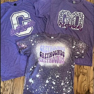 May include: Three purple t-shirts with different designs. The first shirt has a white letter "C" with the word "Greyhounds" in white letters. The second shirt has a white "GO" with a leopard print design and the word "Greyhounds" in white letters. The third shirt has a white splatter paint design with the word "Greyhounds" repeated three times in different shades of purple.