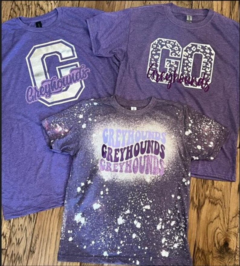 May include: Three purple t-shirts with different designs. The first shirt has a white letter "C" with the word "Greyhounds" in white letters. The second shirt has a white "GO" with a leopard print design and the word "Greyhounds" in white letters. The third shirt has a white splatter paint design with the word "Greyhounds" repeated three times in different shades of purple.