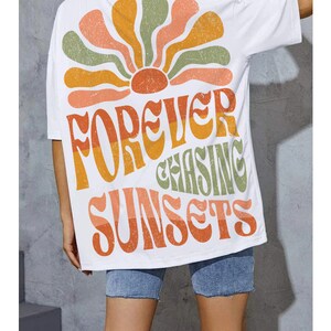 May include: White t-shirt with a retro-style design featuring a stylized sunburst in orange, yellow, and green. The shirt has the text "FOREVER CHASING SUNSETS" in a matching retro font. The shirt is worn with denim shorts.