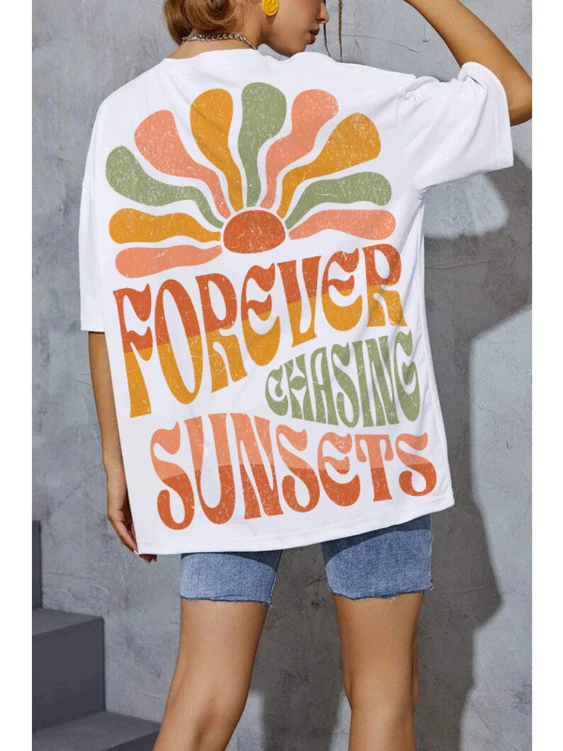 May include: White t-shirt with a retro-style design featuring a stylized sunburst in orange, yellow, and green. The shirt has the text "FOREVER CHASING SUNSETS" in a matching retro font. The shirt is worn with denim shorts.