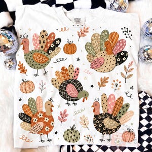 Può includere: Una t-shirt bianca con una stampa grafica ripetuta di cinque tacchini con piume decorate in tonalità arancione, marrone, rosa e nero, intervallati da piccole zucche arancioni, foglie autunnali e stelle. L'etichetta sul collo riporta Comfort Colors.