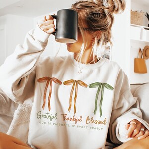 May include: Cream-colored sweatshirt with three ribbon graphics in shades of brown and green. The text "Grateful Thankful Blessed" is printed below the ribbons, with the phrase "GOD IS FAITHFUL IN EVERY SEASON". A black mug is held up to the face.
