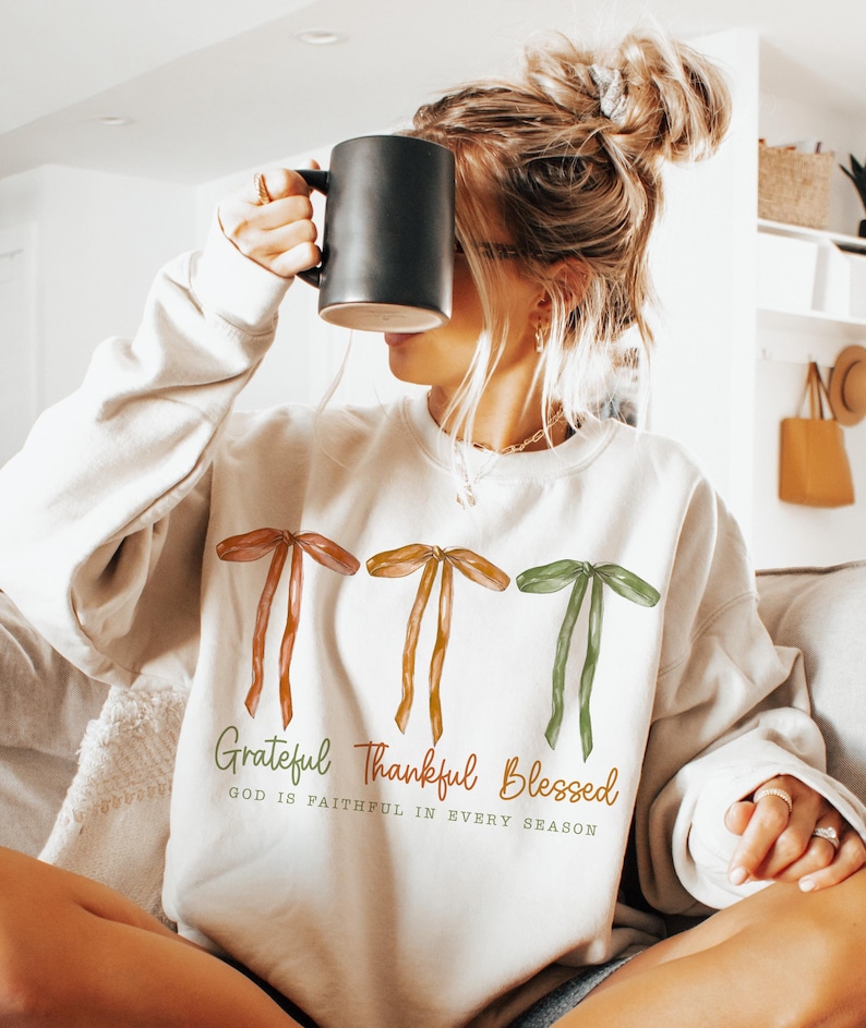 May include: Cream-colored sweatshirt with three ribbon graphics in shades of brown and green. The text "Grateful Thankful Blessed" is printed below the ribbons, with the phrase "GOD IS FAITHFUL IN EVERY SEASON". A black mug is held up to the face.