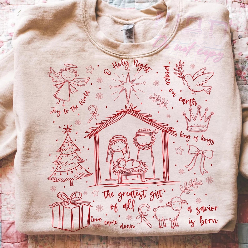 K&ouml;nnte beinhalten: Beiges Sweatshirt mit roter Weihnachts-Grafik. Das Design umfasst eine Krippenszene, einen Engel, einen Stern, einen Weihnachtsbaum, ein Geschenk, ein Schaf und den Text "Holy Night", "Joy to the World" und "Peace on Earth".