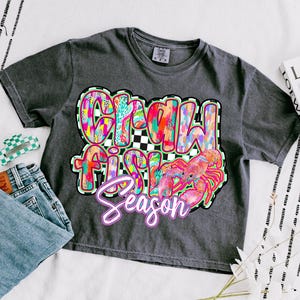 May include: A dark gray t-shirt with a colorful graphic design that says "Crawfish Season" with a cartoon crawfish.