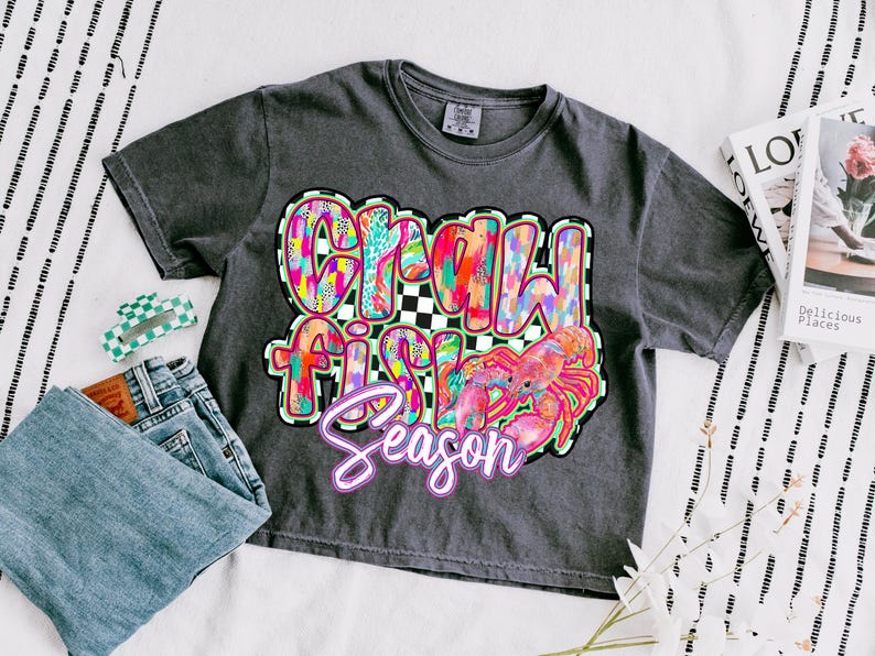 May include: A dark gray t-shirt with a colorful graphic design that says "Crawfish Season" with a cartoon crawfish.