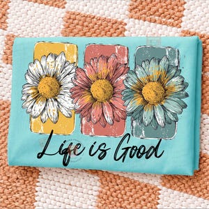 May include: A light blue t-shirt featuring three daisy flowers in yellow, pink, and blue, each set against a rectangular background. The words "Life is Good" are written in black script below the flowers.