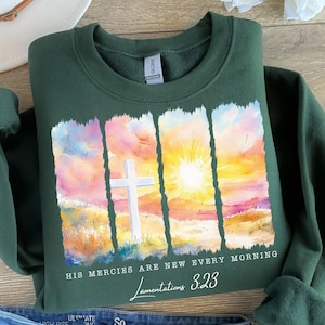 Puede incluir: Sudadera verde con una pintura de acuarela de una salida del sol, una cruz y el texto "Sus misericordias son nuevas cada mañana, Lamentaciones 3:23".