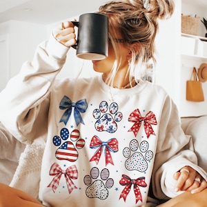 May include: White sweatshirt with a red, white, and blue paw print design. The paw prints are decorated with stars, bows, and polka dots.