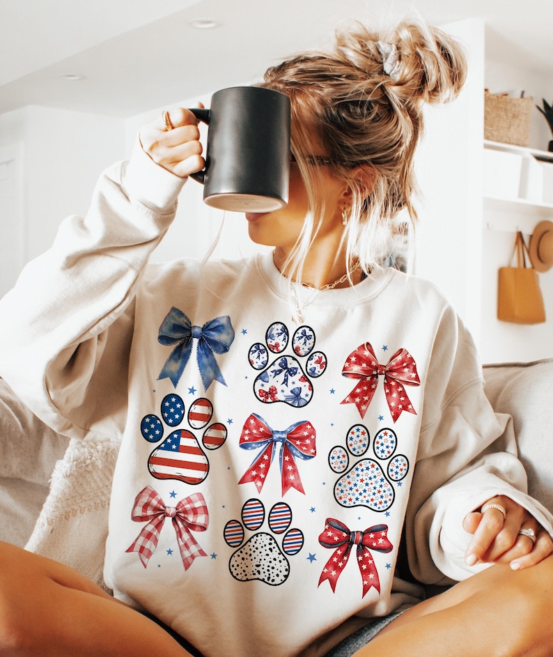 May include: White sweatshirt with a red, white, and blue paw print design. The paw prints are decorated with stars, bows, and polka dots.