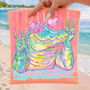 May include: A colorful beach towel featuring a cartoon alligator wearing heart-shaped sunglasses and making peace signs. The alligator is depicted in vibrant shades of blue, green, yellow, and pink against a peach background with pink stripes.