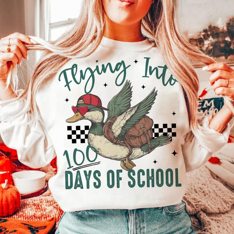 May include: White sweatshirt featuring a green duck wearing a red hat and sunglasses, carrying a brown backpack. The duck is flying with its wings spread. The text "Flying Into 100 Days of School" is printed on the sweatshirt.