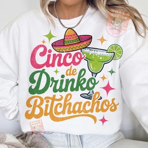 May include: White sweatshirt with a colorful graphic design. The design includes a sombrero, margarita, and the text "Cinco de Drinko Bitchachos" in pink, green, and orange. The design is accented with star shapes.
