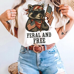 以下が含まれることがあります： サングラス、バンダナ、アメリカ国旗を身に着けたアライグマが描かれたクリーム色のTシャツ。「FERAL AND FREE」の文字が黒い太字で添えられています。このシャツは、青いデニムショーツと茶色のベルトと組み合わせています。