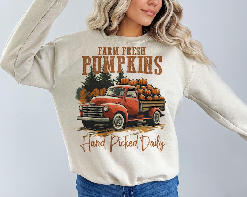 Op de afbeelding: Een beige sweatshirt met een afbeelding van een rode vrachtwagen die pompoenen vervoert. De tekst "Farm Fresh Pumpkins" en "Hand Picked Daily" staat op het shirt gedrukt.